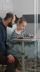 Father sitting with sick daughter in hospital ward while playing online therapy videogames using laptop computer during recovery examination. Girl patient with oxygen nasal tube relaxing after surgery