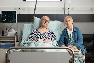 Portrait of grandparents looking into camera during clinical recovery in hospital ward, healthcare...