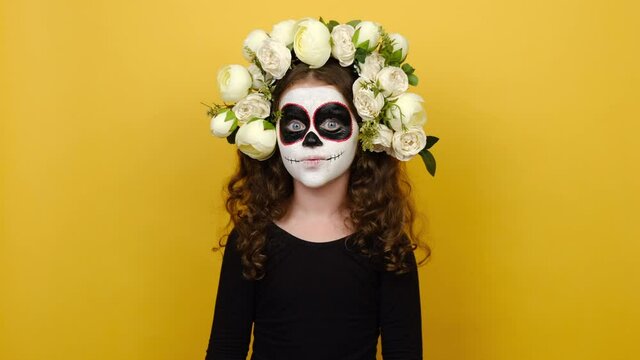 Horrified girl kid with painted skull on face, dressed in black outfit and flowers wreath during day of death to honour dead people, celebrates Mexican version of Halloween, isolated on yellow wall