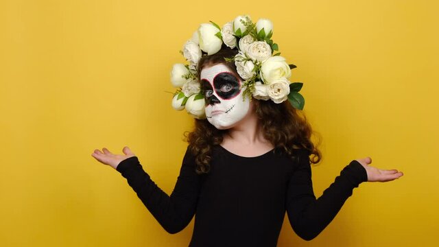 Portrait little girl with Halloween makeup mask wears black outfit and flowers wreath looks around thinks comes, shrugging his shoulders, isolated on yellow wall. Mexican culture, Day of Death concept