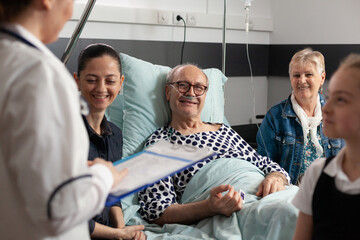 Physician woman doctor discussing healthcare treatment with elderly sick grandfather in hospital ward room. Cheerful caring family supporting retired senior man during clinical recovery