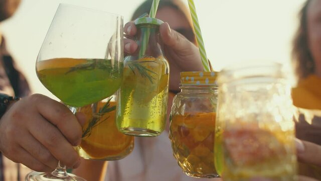 Hands clink glasses with cocktail closeup against background of people in field. Concept of party and fun pastime