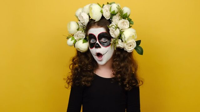 Little girl with Halloween or day of death makeup mask wears black outfit and flowers wreath, looking around with shocked face, isolated on yellow background studio. Celebration holiday party concept