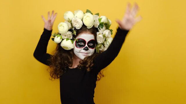 Scary girl kid with Halloween makeup mask, has clay skull face, wears black outfit and flowers wreath dance gesticulating hands fool around have fun enjoy celebrate isolated on yellow background