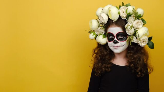 Little girl kid with Halloween makeup mask wears black outfit and flowers wreath looking camera wink eye blink, posing isolated over yellow background in studio. Celebration holiday party concept