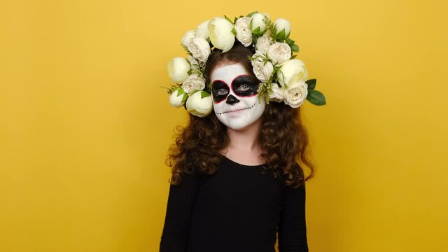 Portrait of schoolgirl kid wears face art mask wears black outfit and flowers wreath point index finger camera on you, posing isolated on yellow studio background. Day dead, Happy Halloween concept
