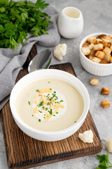 Homemade cauliflower soup puree in a white bowl with cheese, cream, spices and fresh parsley on a gray concrete background. Top view, copy space.