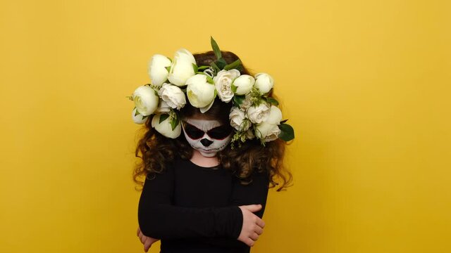 Angry sad little girl with halloween makeup, has clay skull face, wears black outfit and flowers wreath, crossed arms looking to side, isolated on yellow studio background. Day of Death concept