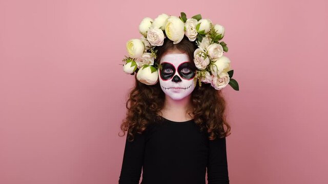 Secret bright girl kid with Halloween makeup mask wears black outfit and flowers wreath, looking camera close her mouth lock, isolated on pink studio background. Mexican culture, Day of Death concept