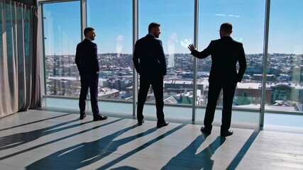 Serious businessmen standing near the windows. Elegant entrepreneurs in suits listen to their partner indoor the skyscraper. Back view.