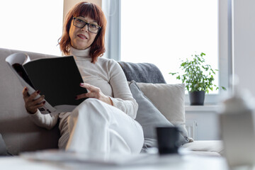 middle-aged adult woman with glasses sits on the sofa and reads a magazine