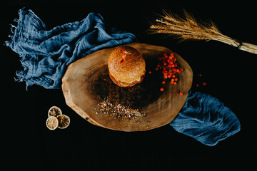 Hamburger on wooden plate with small grains of spice, wheat, red currant and dried lemon on black background and blue shawl