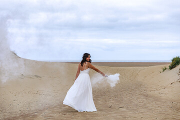 Fototapeta na wymiar Junge schöne Frau im Hochzeitskleid am Strand mit einer Rauchfackel und weißen Rauch