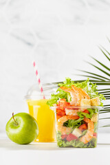 Healthy summer vacation daily diet plan - set of orange juice, green apple, fresh tropical salad with shrimps and vegetables in plastic box  in white interior with palm leaf, vertical.