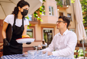 Waitress in protective mask serving delicious food for man in cozy restaurant. Coronavirus outbreak lifestyle. Social distancing. COVID-19.