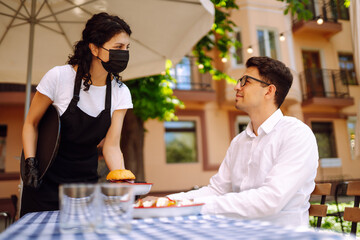 Waitress in protective mask serving delicious food for man in cozy restaurant. Coronavirus outbreak lifestyle. Social distancing. COVID-19.