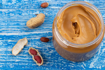 Peanut butter in an open jar and peanuts in the skin are scattered on the blue table. Space for...