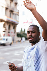 young afro man standing on the street and raising arm to call to attention of a cab. He is holding...