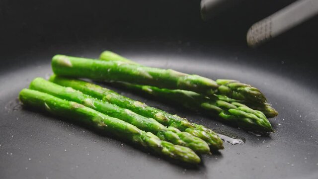 green asparagus is fried in a Frying pan with a Teflon coating. tongs turn over the greens.