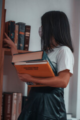 Tutora con mascarilla eligiendo libros de la estanteria de clases para que sus alumnos estudiantes...