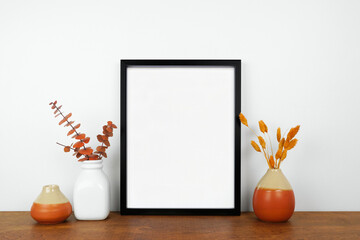Mock up black frame with fall home decor on a wood shelf. Autumn concept. Portrait frame against a...