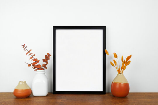 Mock up black frame with fall home decor on a wood shelf. Autumn concept. Portrait frame against a white wall.