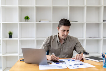 Fototapeta premium Young businessman sitting and checking financial documents displayed in chart format, young businessman who founded a startup company, managing a new business to grow by leaps and bounds.