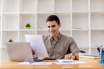 Fototapeta premium Young businessman sitting in office with laptop, he is reading company financial briefs, he founded a startup, he is a young entrepreneur who is skilled in running a growing company.