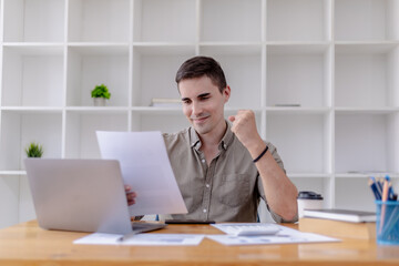 Fototapeta premium Young businessman sits in his office with a laptop, he looks delighted as he reads the documents that the company is profitable, he is a young businessman who is skilled in running the company to grow