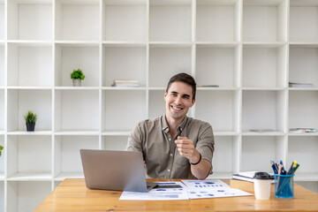 Fototapeta premium Young businessman sitting in the office working with laptop and paperwork on the desk, smiling, he is the founder of a startup, he is a young businessman who is skilled in running a growing company.