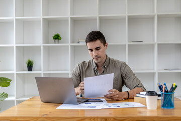 Fototapeta premium A young businessman sitting in the office working with a laptop, he is checking financial documents, he founded a startup, he is a young entrepreneur who has the skills to run a growing company.