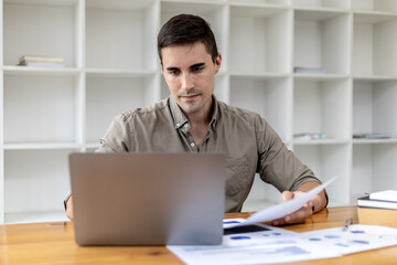A young businessman sitting in the office working with a laptop, he is checking financial documents, he founded a startup, he is a young entrepreneur who has the skills to run a growing company.