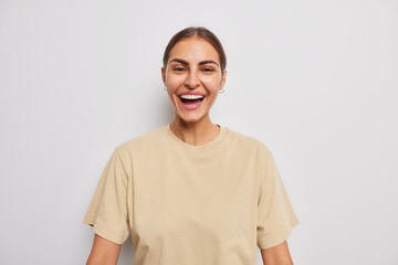 Carefree cheerful woman with dark combed hair smiles toothily shows white teeth dressed in casual beige t shirt poses against grey background expresses positive emotions pleased with results