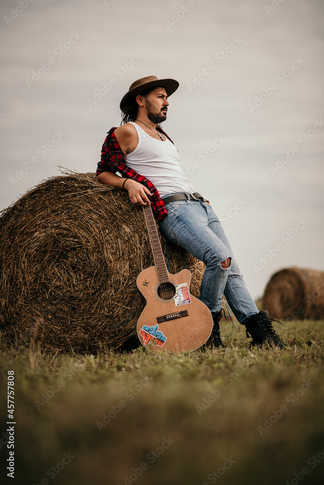 Poster Young handsome Serbian male in a flannel shirt leaned on a haystack holding his guitar