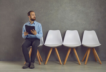 Stressed caucasian man feeling fear before job interview sitting on chair holding clipboard with CV...