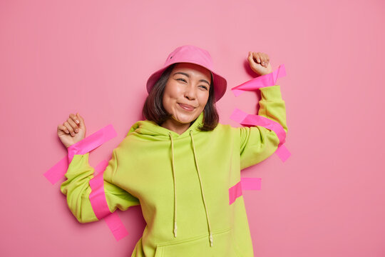 Happy attractive brunette Asian woman stuck to walll with plaster dressed in casual clothes looks with dreamy thoughtful expression forward recalls pleasant event isolated over pink background