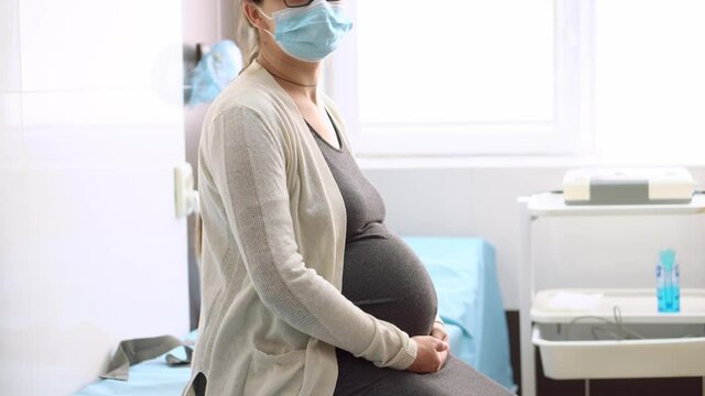 Young Pregnant Woman In Medicine Protective Mask On Hospital Couch Before Medical Control Cardiotocography. Visit Gynecologist Doctor At Clinic. Examine Expectant Belly Baby Mother Healthcare Check Up