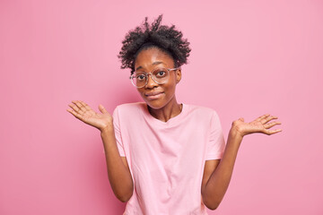 Young dark skinned woman shrugs shoulders shows empty hands has no clue about something nothing to say stands unaware indoor wears big optical glasses casual t shirt isolated over pink wall.