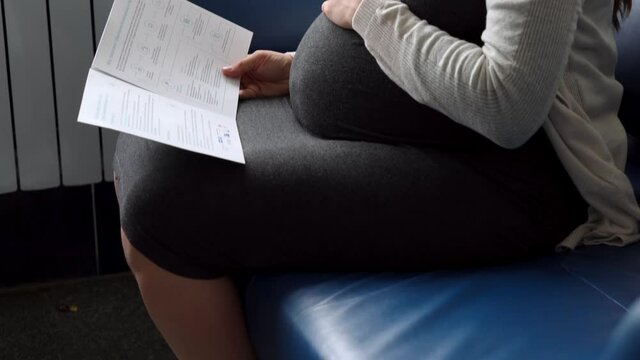 Young Pregnant Woman In Medicine Protective Mask Reading On Couch Waiting For Appointment Medical Control On Hospital. Visit Gynecologist Doctor At Clinic. Examine Expectant Belly Healthcare Check Up