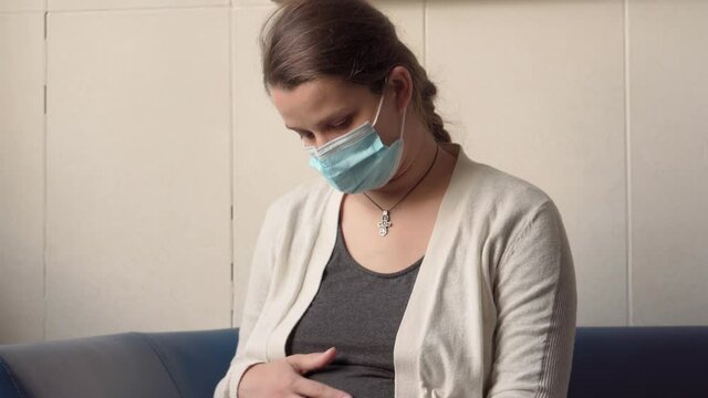 Young Pregnant Woman In Medicine Protective Mask Reading On Couch Waiting For Appointment Medical Control On Hospital. Visit Gynecologist Doctor At Clinic. Examine Expectant Belly Healthcare Check Up