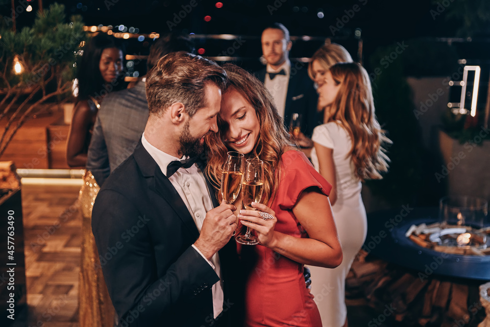 Sticker beautiful young couple in formalwear holding champagne flutes and smiling while spending time on lux