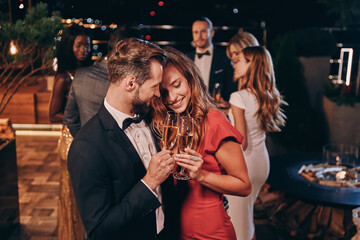 Beautiful young couple in formalwear holding champagne flutes and smiling while spending time on luxury party