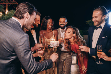 Beautiful young woman in evening gown blowing candles on cake while celebrating birthday with...