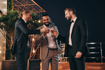 Three handsome men in suits drinking whiskey and communicating while spending time on party