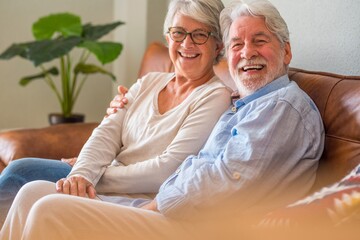 Portrait of cheerful senior couple embracing while sitting on sofa and smiling. Elderly happy couple relaxing and posing in front of camera sitting in living room.