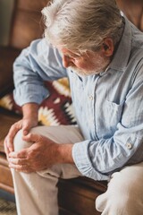 Senior old man facing knee problem, sitting on sofa holding knee at home. Elderly man suffering...
