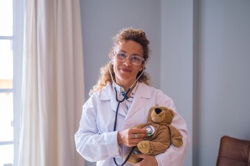 Portriat of young female doctor in uniform checking teddy bear with stethoscope at clinic. Confident medical practicitioner or healthcare worker examining teddy bear at hospital