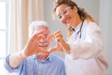 Close up of young female doctor and senior man showing heart sign with hand at home. Doctor helping...