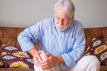 Senior old man facing knee problem, sitting on sofa holding knee at home. Elderly man suffering...