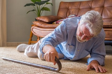 Elder senior man lying on floor after falling down with wooden walking stick beside couch on rug in...
